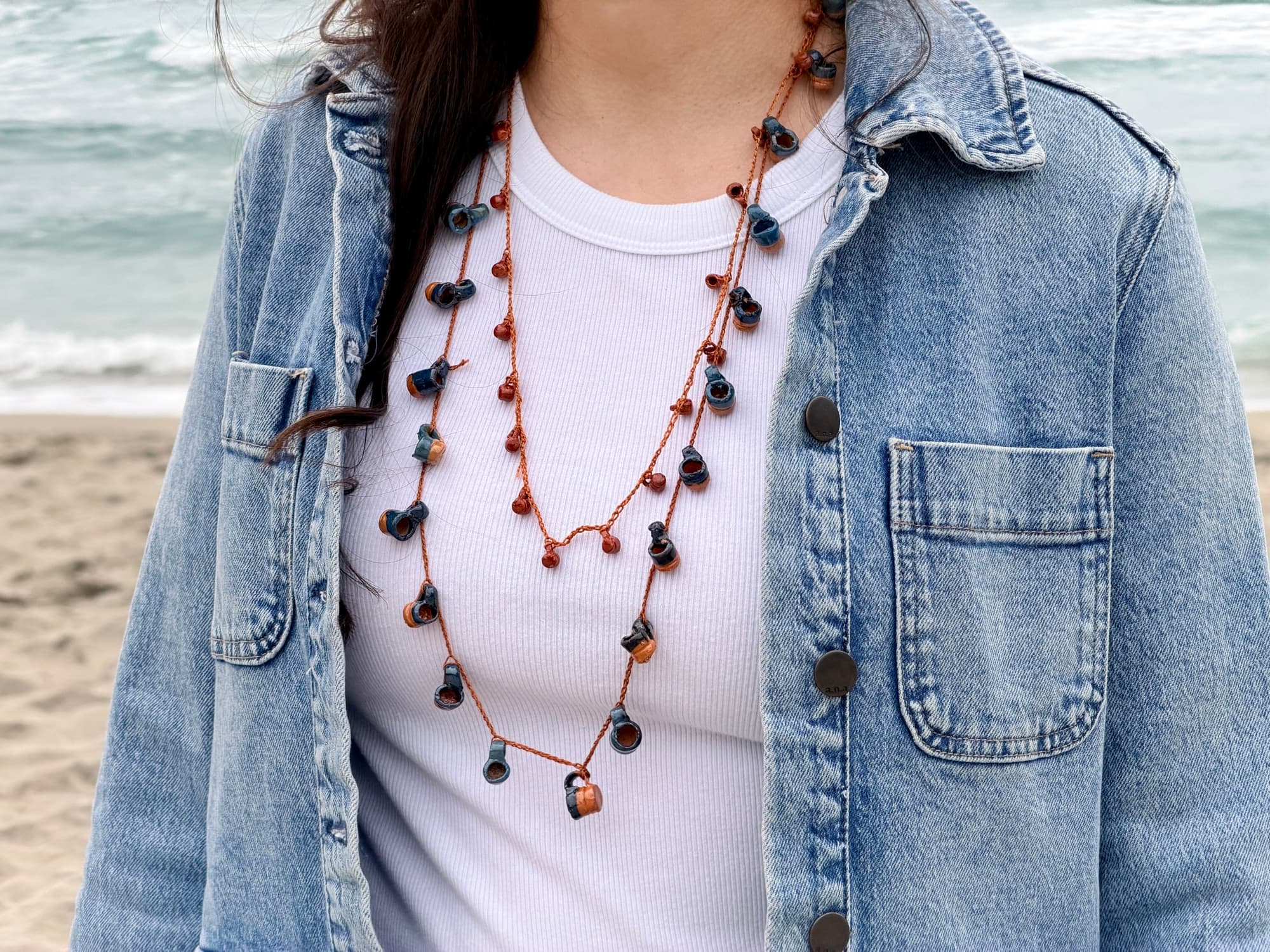 Blue and terracotta jarrito collars layered over a white top and denim jacket by the ocean.