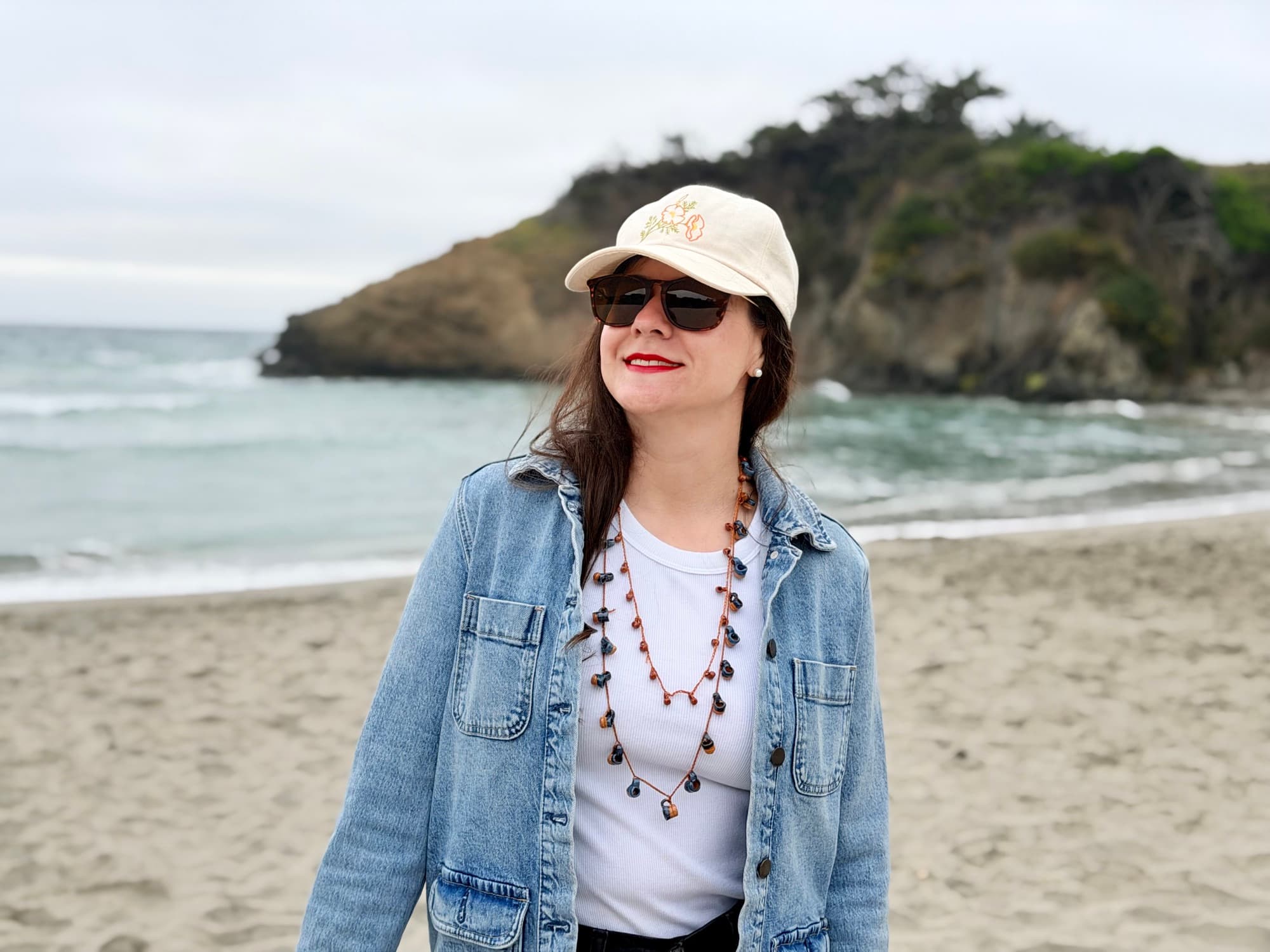 A woman smiling on the beach in a denim jacket, wearing two handmade jarrito collars layered together.